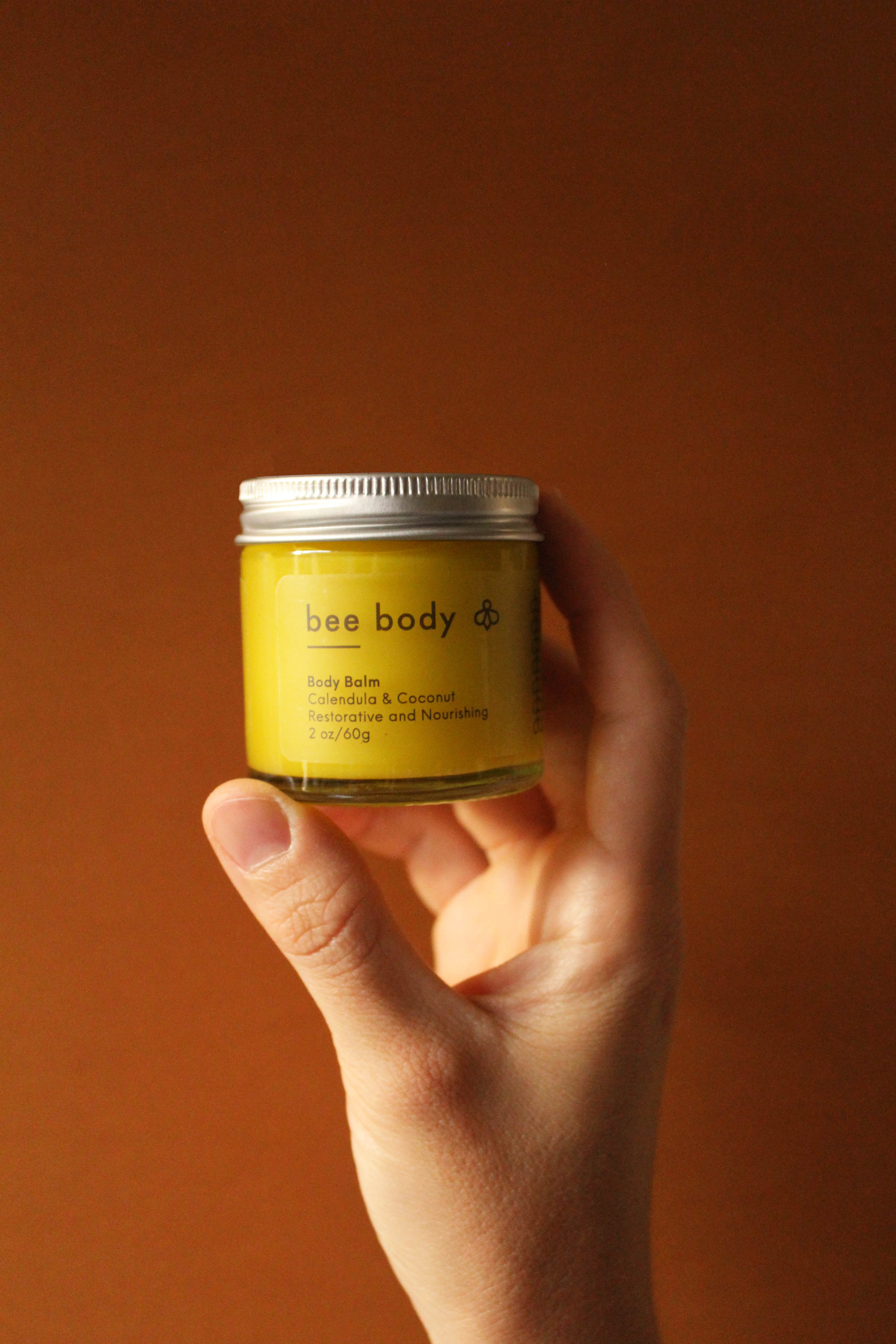 Calendula & Coconut Body Balm: Elegantly held in hand against warm honey-coloured backdrop, experience the soothing blend of calendula and coconut for nourished skin. 