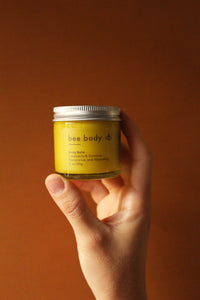 Calendula & Coconut Body Balm: Elegantly held in hand against warm honey-coloured backdrop, experience the soothing blend of calendula and coconut for nourished skin. 