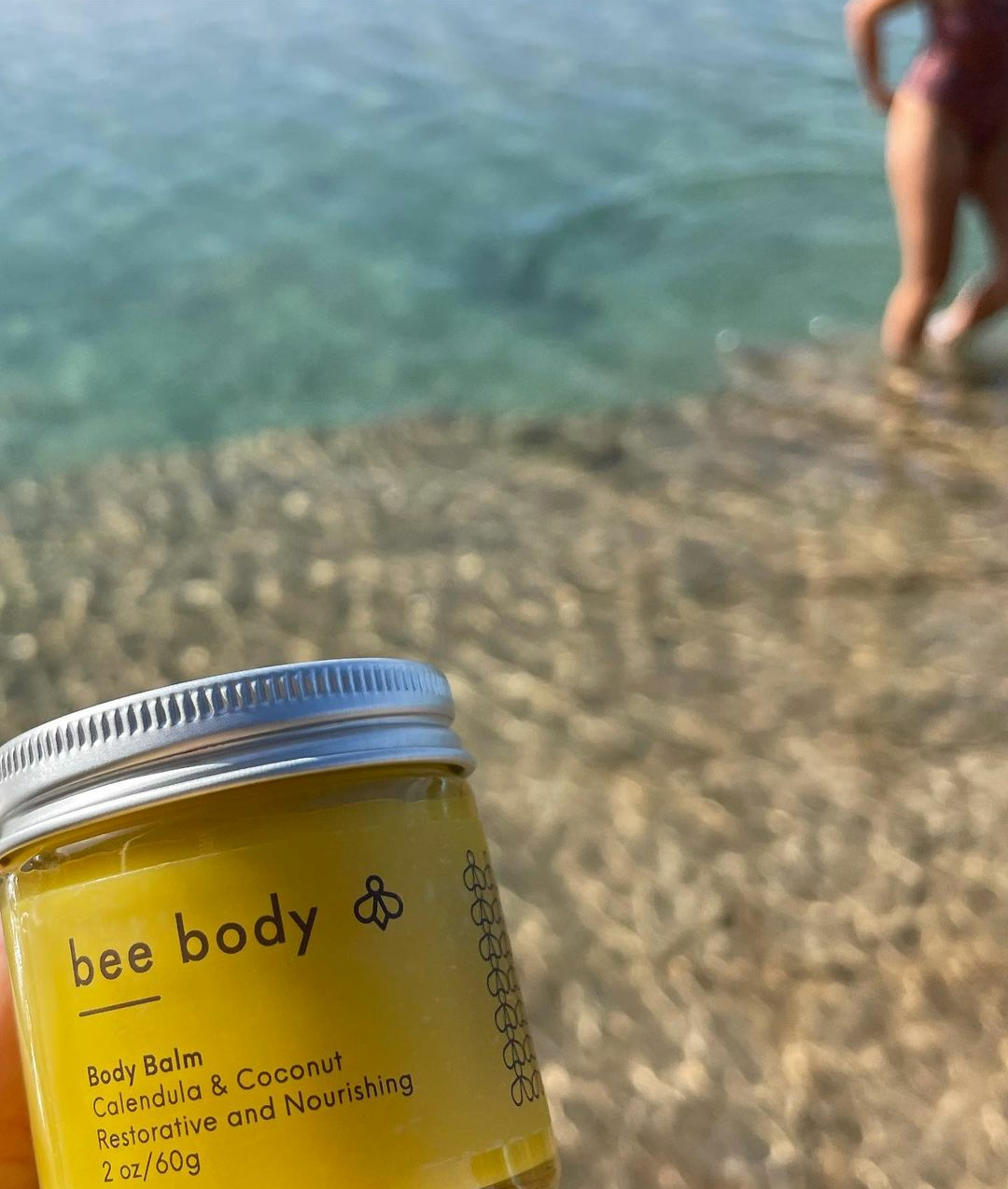 Calendula & Coconut Body Balm: Embrace nature’s soothing embrace as the balm is held amidst a serene beach setting. With pebbles underfoot and turquoise waters glistening, a tranquil backdrop frames the scene, where a relaxed female figure enjoys the beauty of the beach in her bathing suit.