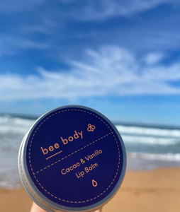 Cacao & Vanilla Lip Balm: Enjoy the nourishing blend of beeswax, honey, cacao and vanilla with the product held against a picturesque Australian beach backdrop. Embrace the soothing benefits under a clear blue sky and scenic natural environment.