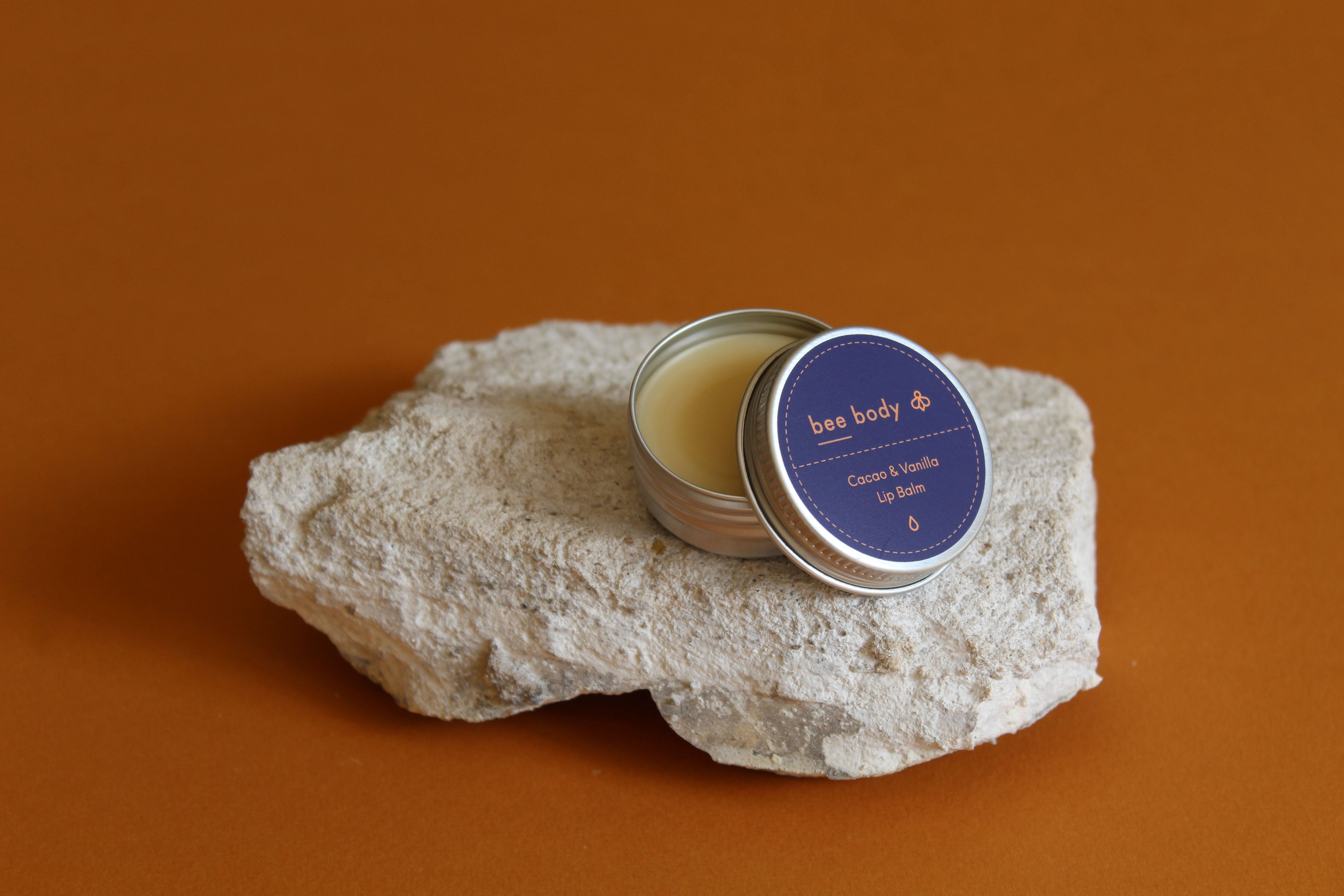 Cacao & Vanilla Lip Balm: Nourishing blend of beeswax, honey, cacao and vanilla resting gracefully on a natural rock with the lid off. Experience the soothing benefits of our product amidst a serene honey-coloured backdrop.