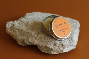 Macadamia & Honey Lip Balm: Discover the essence of nature as the lid is slightly off, revealing the nourishing balm inside, resting gracefully on a natural rock. 