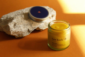 Calendula & Coconut Body Balm: Lid resting on a rock, balm beside it, revealing its golden colour and rich texture for a soothing skin experience. 