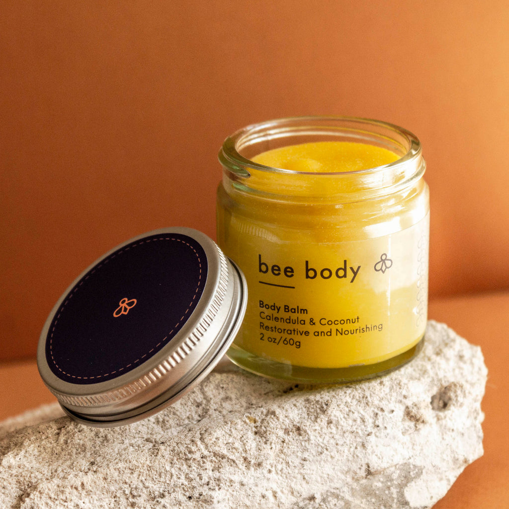 Calendula & Coconut Body Balm: Discover the nourishing blend of calendula, coconut, beeswax and honey in a clear glass jar, with the lid resting gracefully beside it. Set on a light grey rock against a warm honey-coloured backdrop.