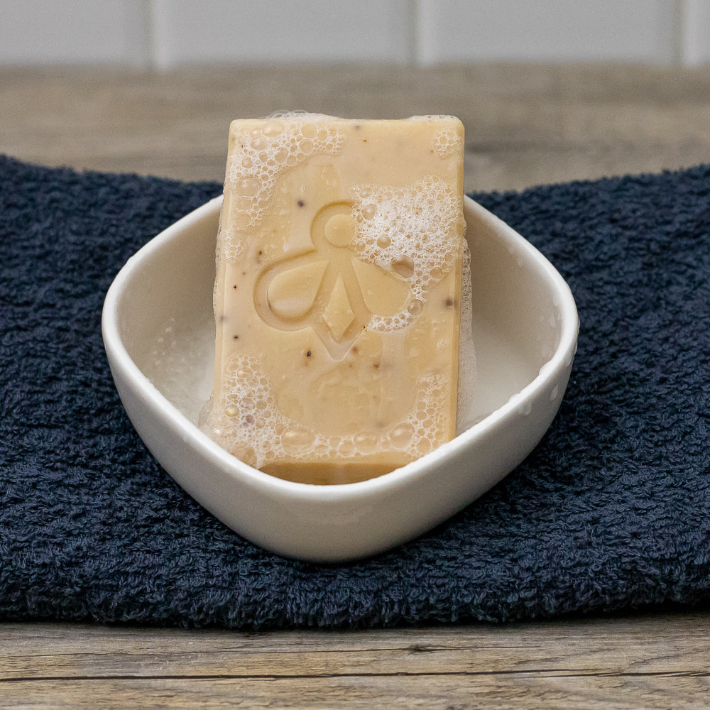 Lavender and Goat’s Milk Soap in Dish - A tranquil scene with our soap resting in a dish, surrounded by delightful suds, all atop a soft navy blue towel. Experience the calming blend of lavender and goat’s milk, designed for a gentle and nourishing bathing experience.