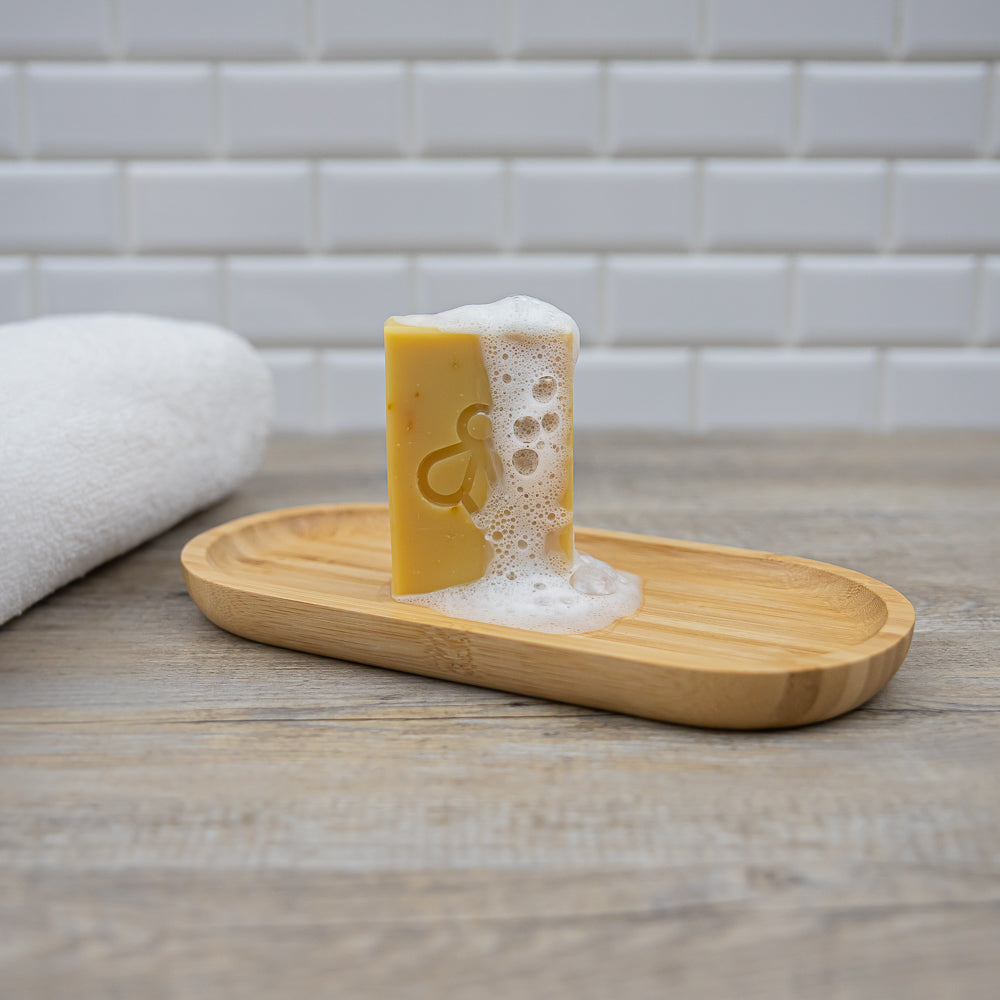 Sunshine Soap on Bamboo Dish in Bathroom - Radiant and invigorating soap with suds, showcased against a backdrop of white tiles, white towel, and wooden benchtop.