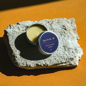 Cacao & Vanilla Lip Balm: A luxurious blend of beeswax, honey, cacao and vanilla lip balm showcased with the lid off. Placed on a rock with honey-coloured paper background. Nourish your lips with our quality lip care product. 