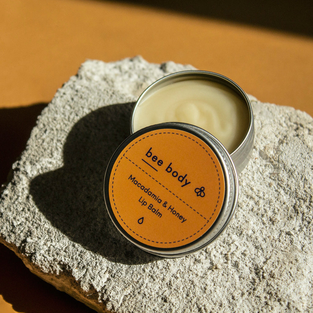 Macadamia & Honey Lip Balm: Experience the natural goodness of macadamia and honey lip balm as it rests gracefully on a rock, the open tin revealing the soothing balm. Nourish your lips amidst the tranquil setting of the rock on a honey-coloured backdrop. 